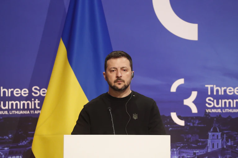 Ukraine's president, Volodymyr Zelensky stands at a podium flanked by the Ukraine flag.