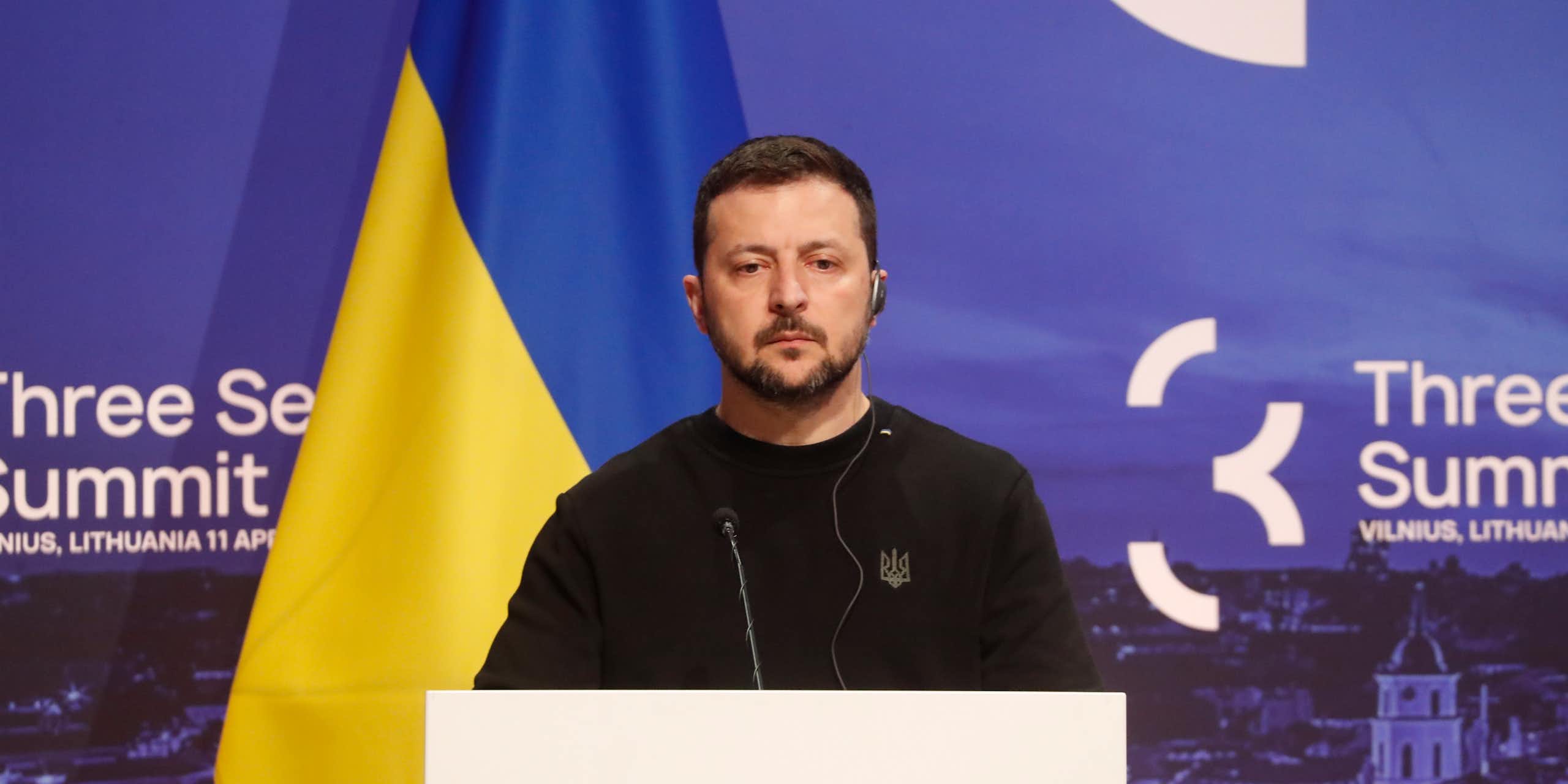 Ukraine's president, Volodymyr Zelensky stands at a podium flanked by the Ukraine flag.