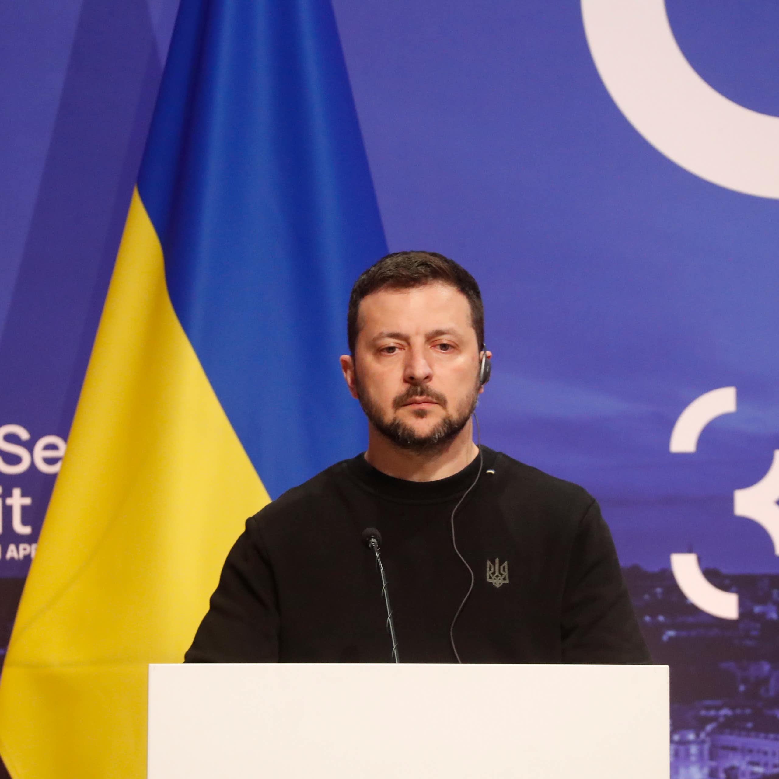 Ukraine's president, Volodymyr Zelensky stands at a podium flanked by the Ukraine flag.