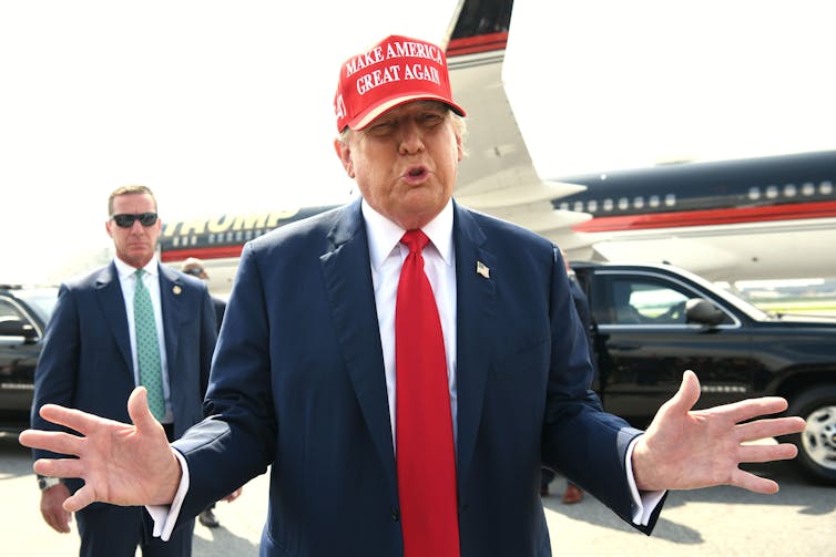 Donaldn Trump in a MAGA hat holds out both hands. In the background are a security guard and his private jet.