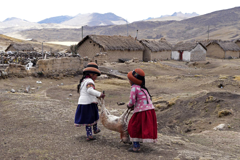 Two girls carry a sheep in front of some buildings.