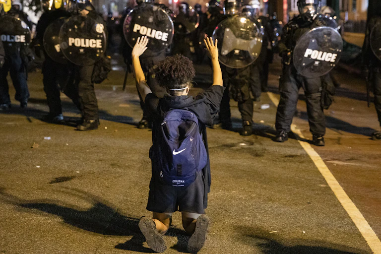 A person wearing a backpack kneels and holds their hands above their head as police in helmets and carrying shields approach.