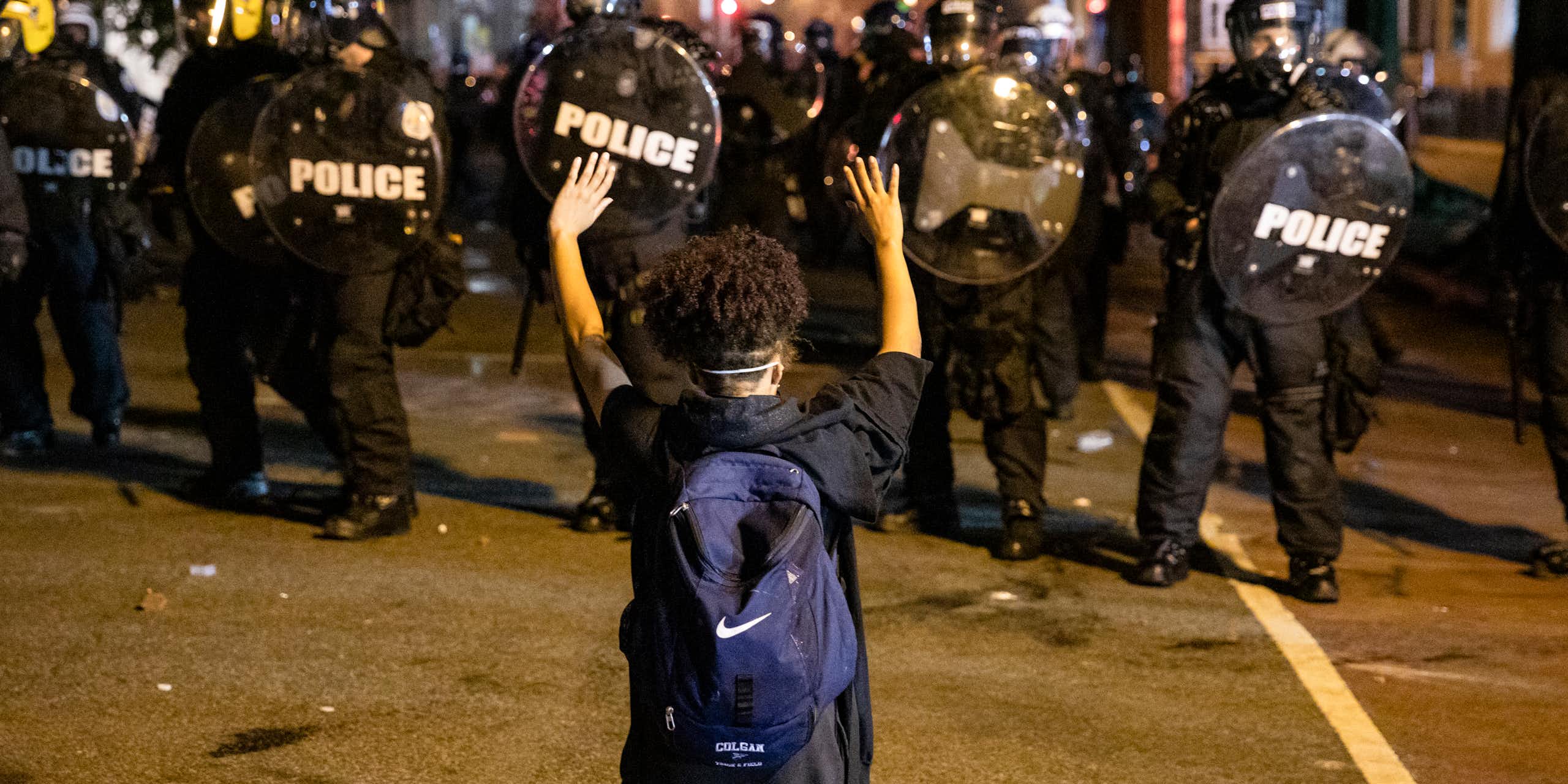 A person wearing a backpack kneels and holds their hands above their head as police in helmets and carrying shields approach.