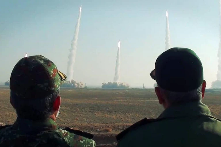 Two men in baseball caps look on as missiles launch surrounded by plumes of smoke.