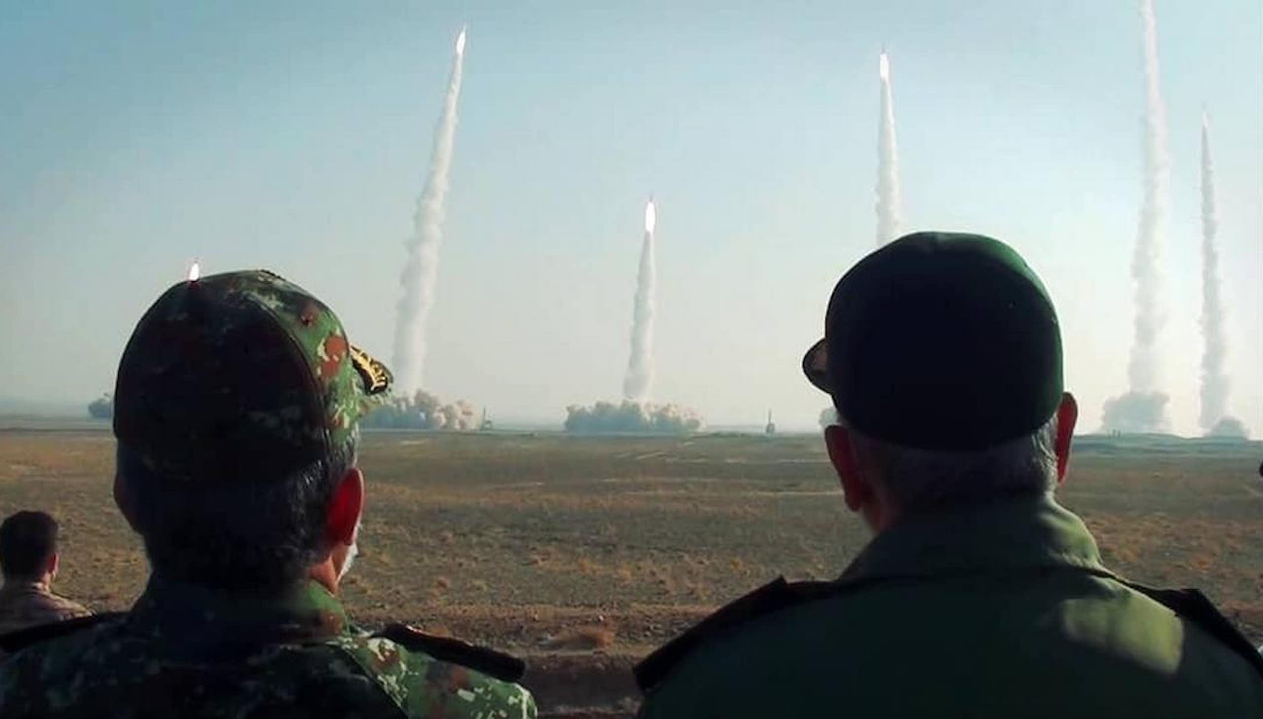 Two men in baseball caps look on as missiles launch surrounded by plumes of smoke.