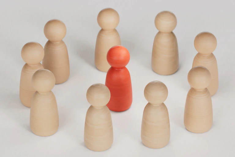 ten figure-like wooden cylinders topped by spheres with nine beige ones arranged in a circle around one red one