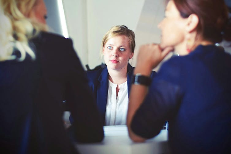 Three woman sit at a table. One looks intently at another who is speaking.