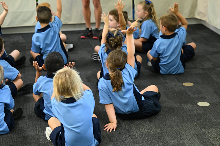 Children in school uniforms raise their hands while sitting on the floor.