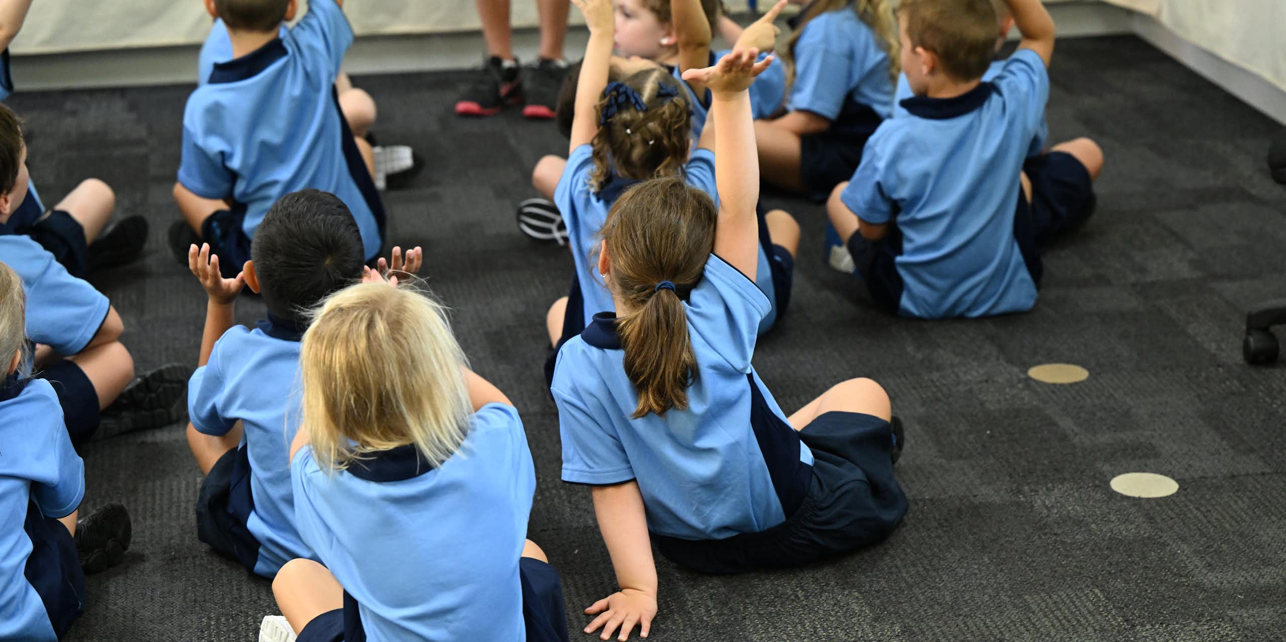 Children in school uniforms raise their hands while sitting on the floor. 