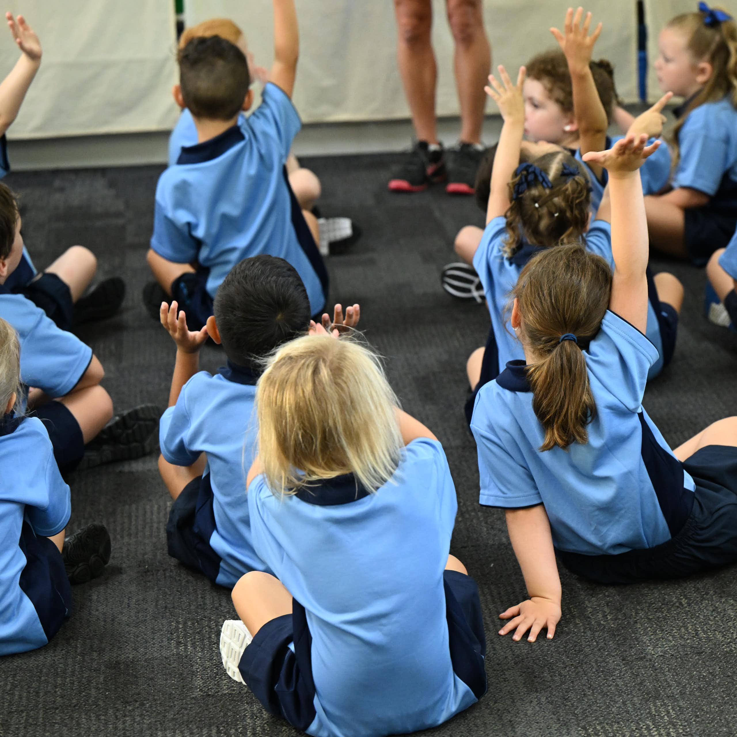 Children in school uniforms raise their hands while sitting on the floor. 