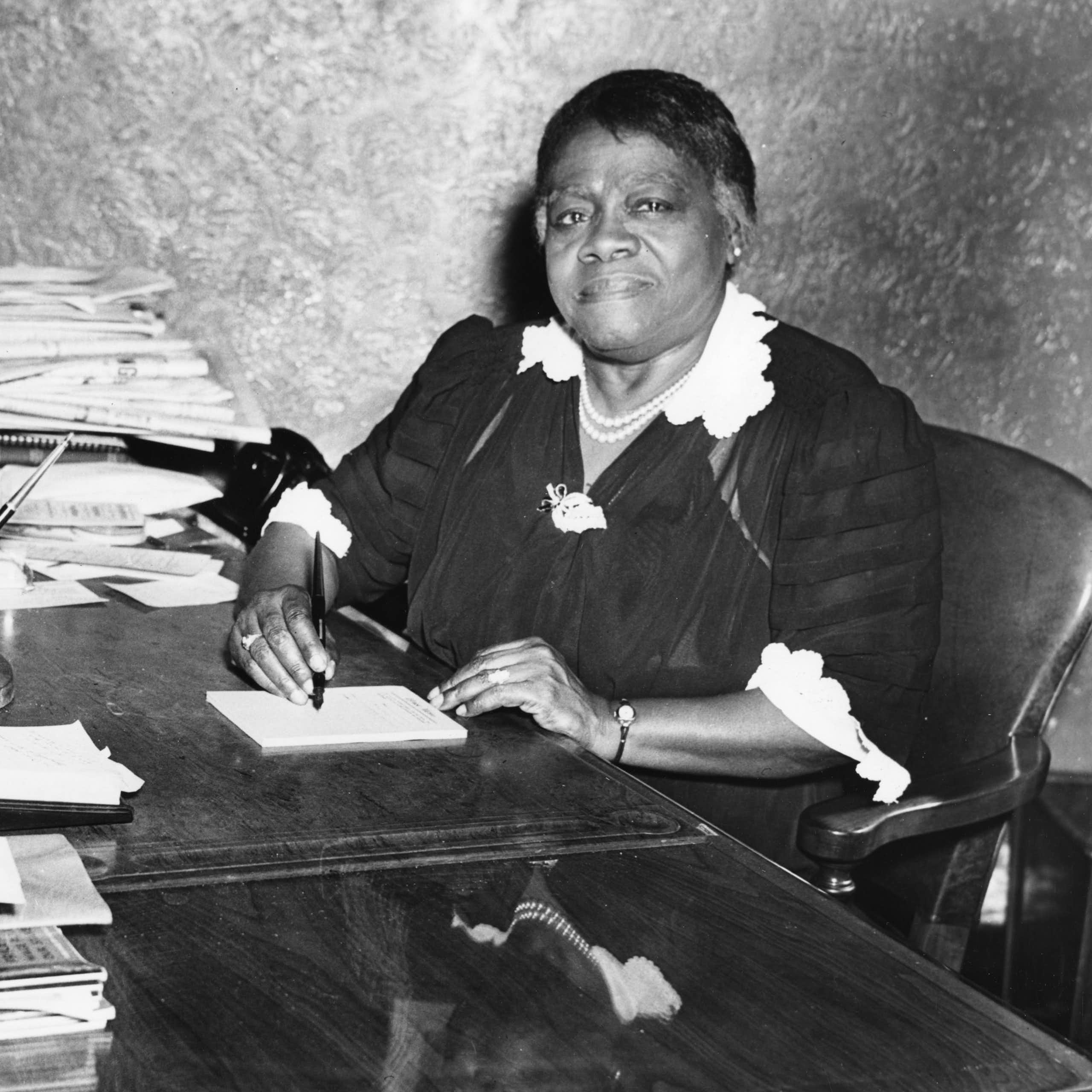 A woman sits at a desk filled with stacks of papers. She has a pen and pad.