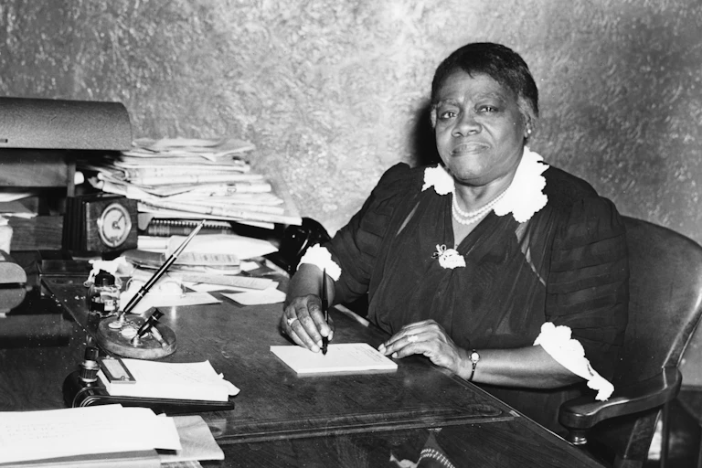 A woman sits at a desk filled with stacks of papers. She has a pen and pad.