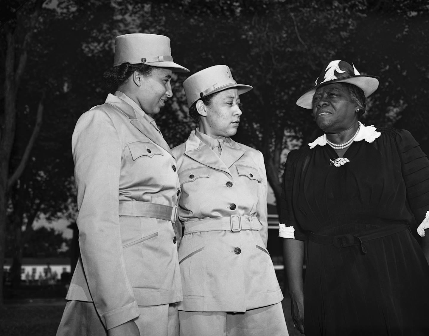 Two women in military uniforms speak to a woman in civilian clothes.