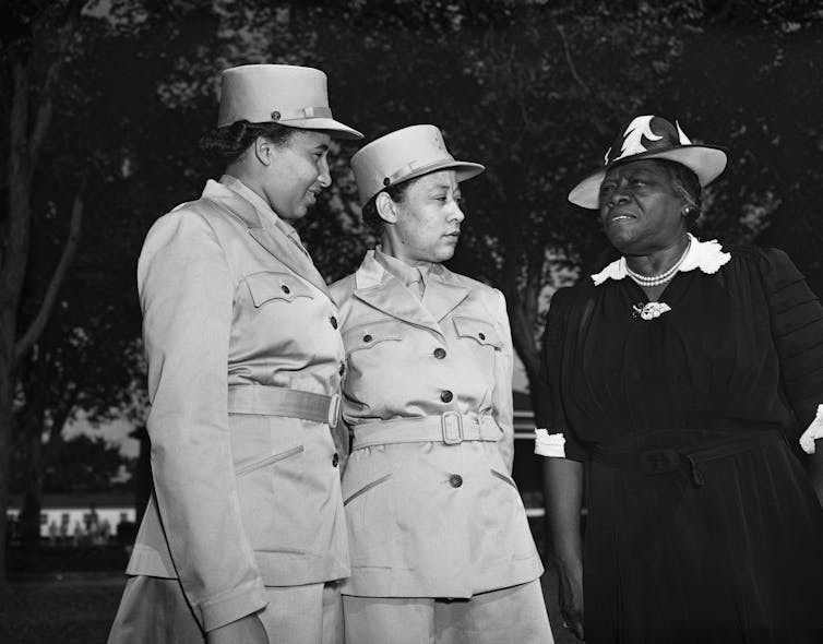 Two women in military uniforms talk to a woman in civilian clothes.