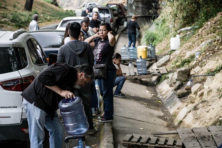 People collecting water from a pipe next to a road.