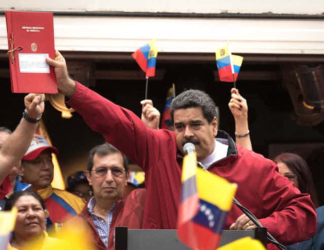 Venezuelan president Nicolas Maduro holding a red book with Venezuela flags waving around him.