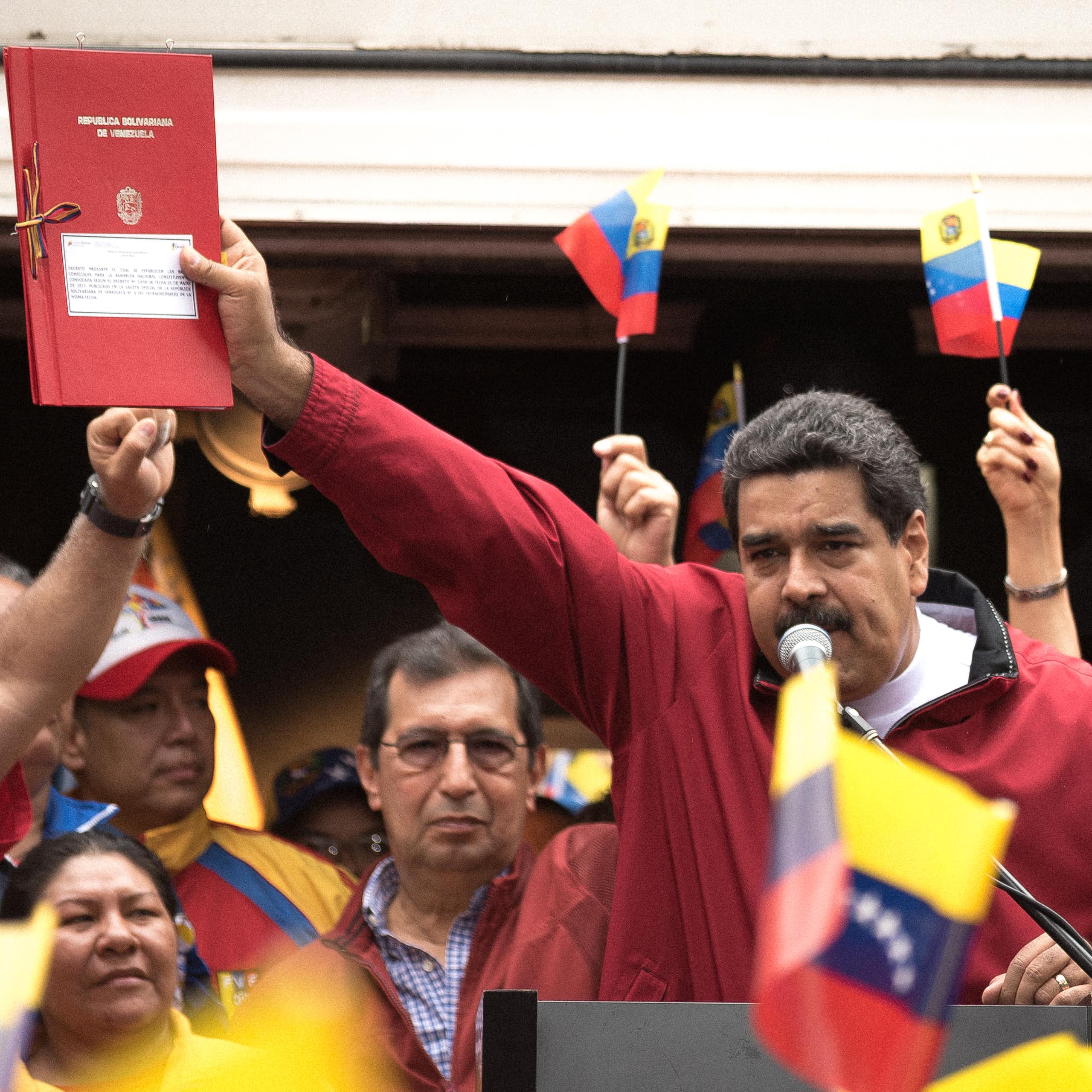 Venezuelan president Nicolas Maduro holding a red book with Venezuela flags waving around him.
