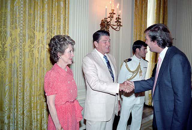 A man in a white suit shakes hands with a younger man in a navy suit while a woman in a pink dress looks on.