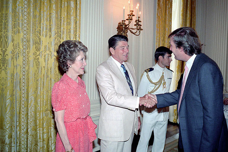 A man in a white suit shakes hands with a younger man in a navy suit while a woman in a pink dress looks on.