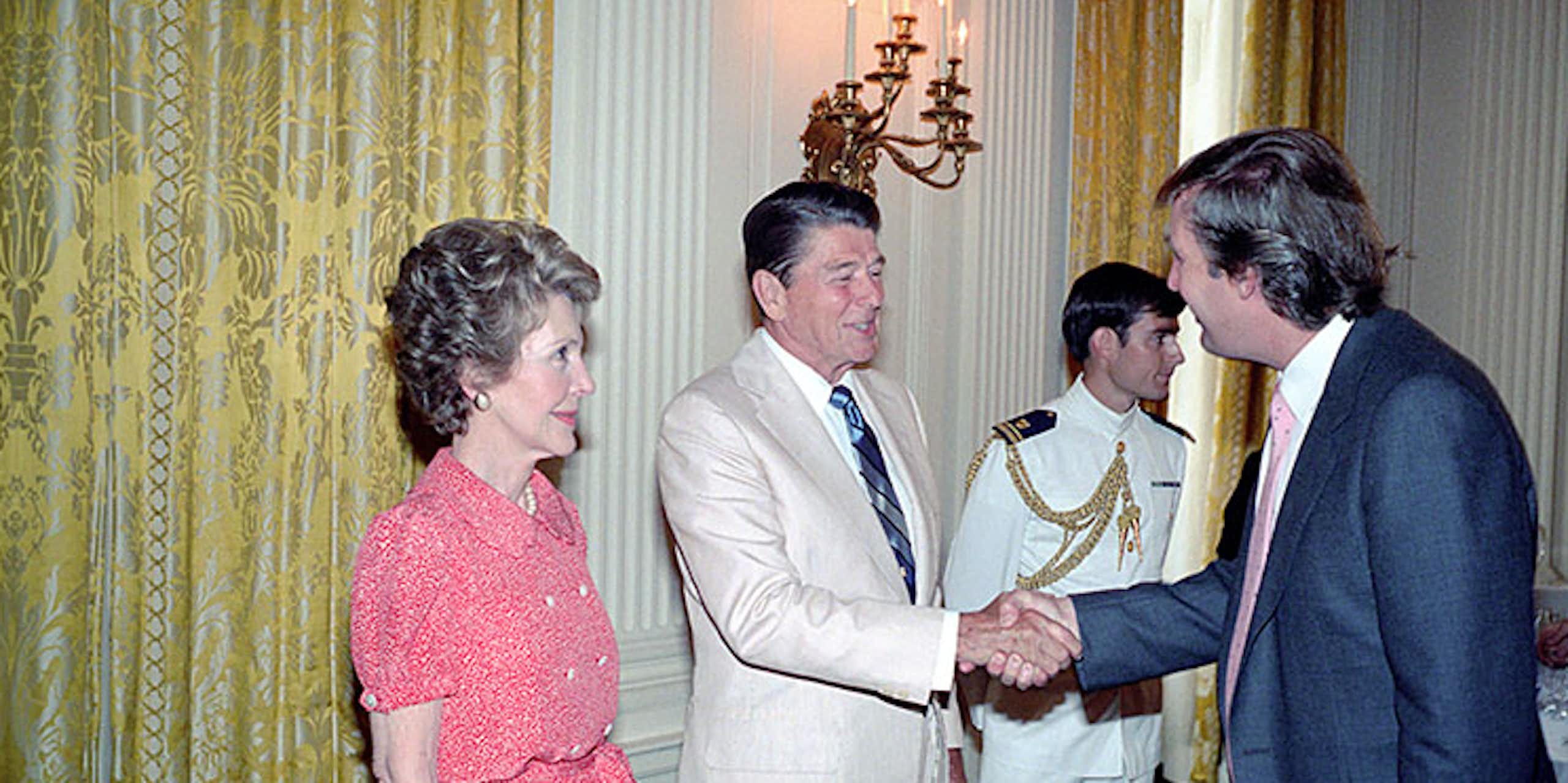 A man in a white suit shakes hands with a younger man in a navy suit while a woman in a pink dress looks on.