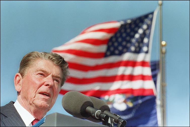 A close up of a man speaking in front of a microphone with the American flag next to him.