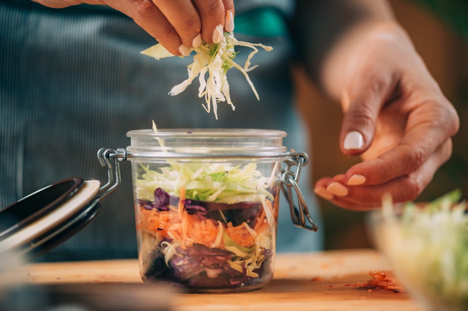 Close-up of person putting shredded cabbage in a jar of other shredded vegetables.
