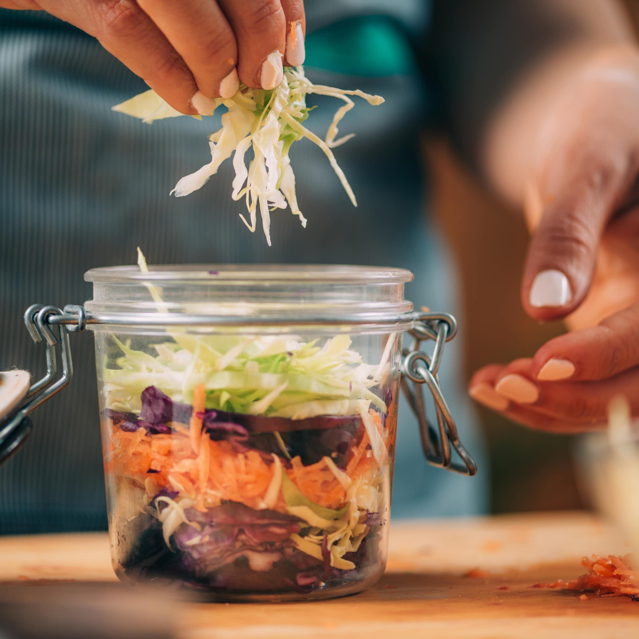 Close-up of person putting shredded cabbage in a jar of other shredded vegetables.