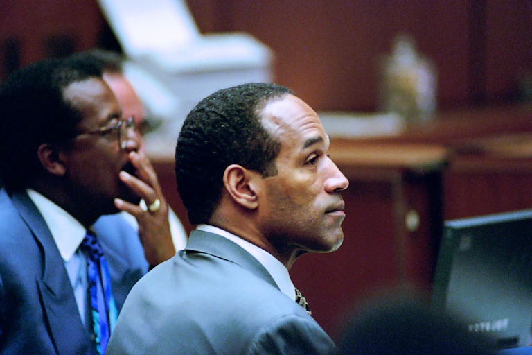 A Black man dressed in a suit sits in a courtroom and is listening to testimony.