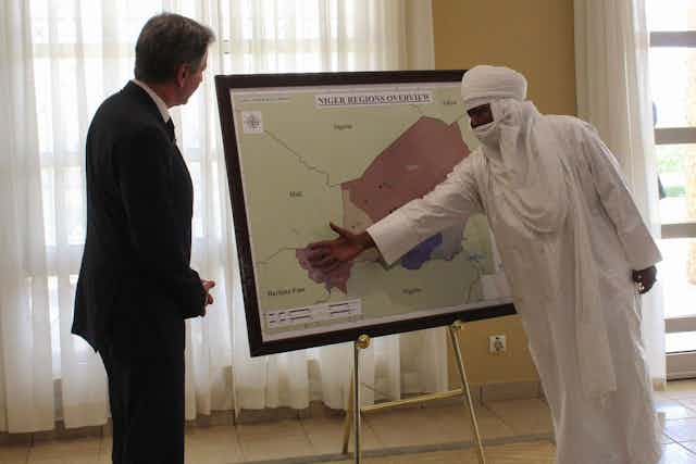 A man covered in white cloth gestures to a map of Niger and the surrounding region while another man in a suit looks on.