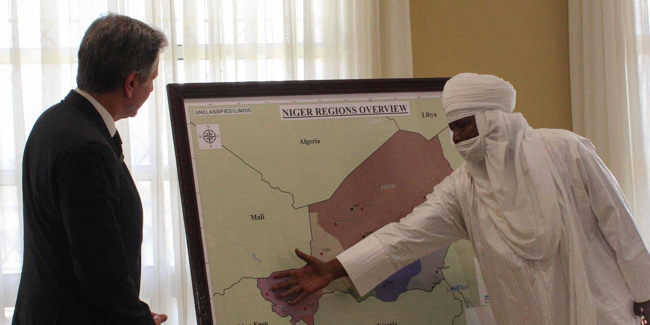 A man covered in white cloth gestures to a map of Niger and the surrounding region while another man in a suit looks on.