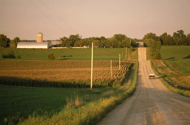 A truck moves past a farm as it drives down a gravel road in a rural area.