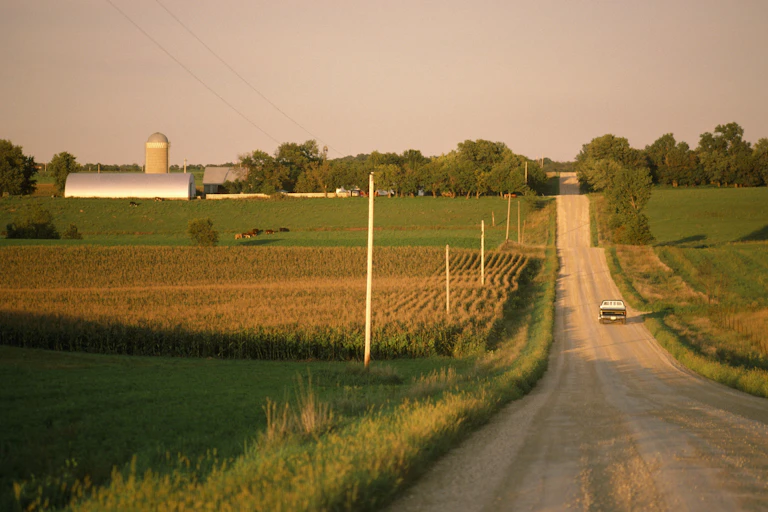 A truck moves past a farm as it drives down a gravel road in a rural area.