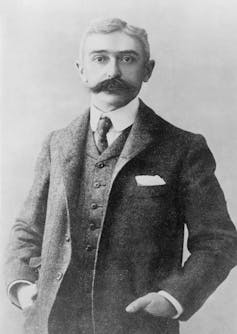 Black and white photo of a man with a moustache and side part standing with his hands in his coat pockets