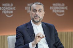 A bearded man in a blue jacket and white collarless shirt gestures as he speaks.