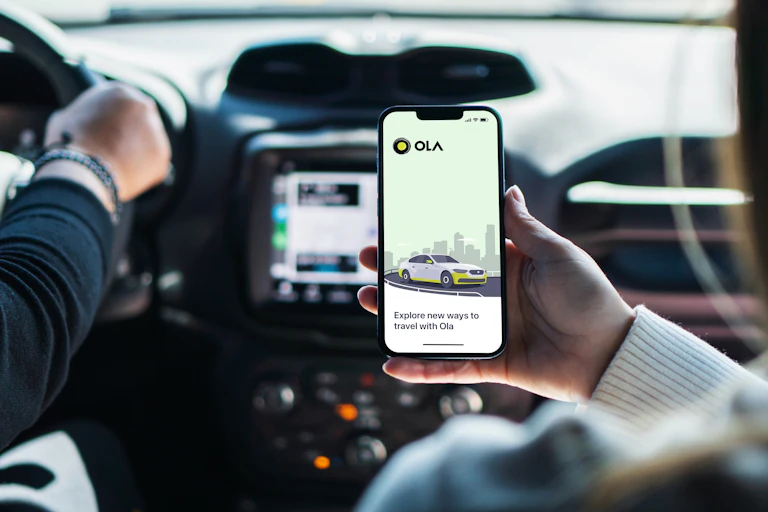Girl inside car holding smartphone with Ola Cabs app on screen.