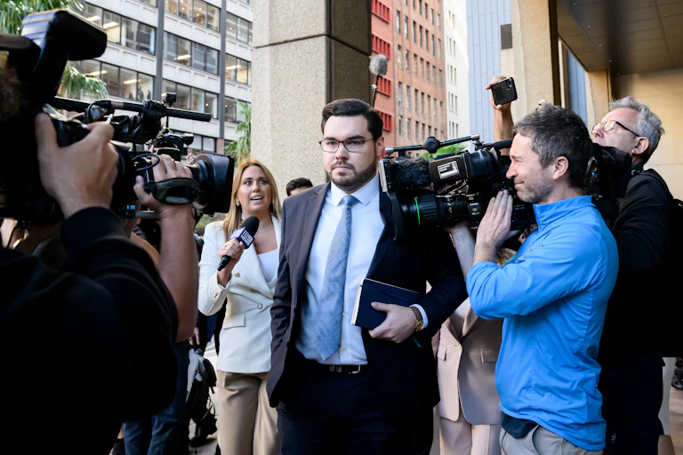 A man in glasses and a suit and tie enters a courtroom amid a crowd of cameras and journalists