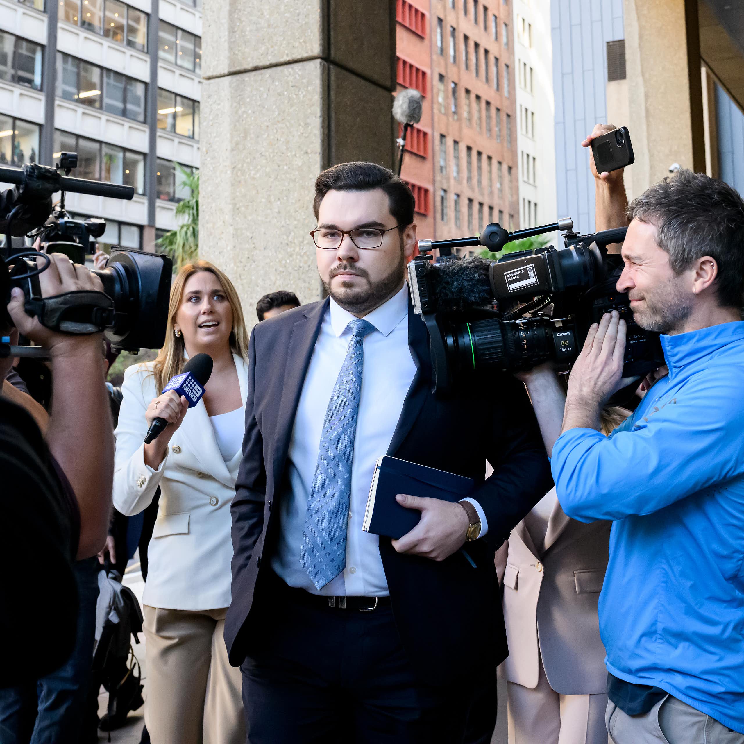 A man in glasses and a suit and tie enters a courtroom amid a crowd of cameras and journalists