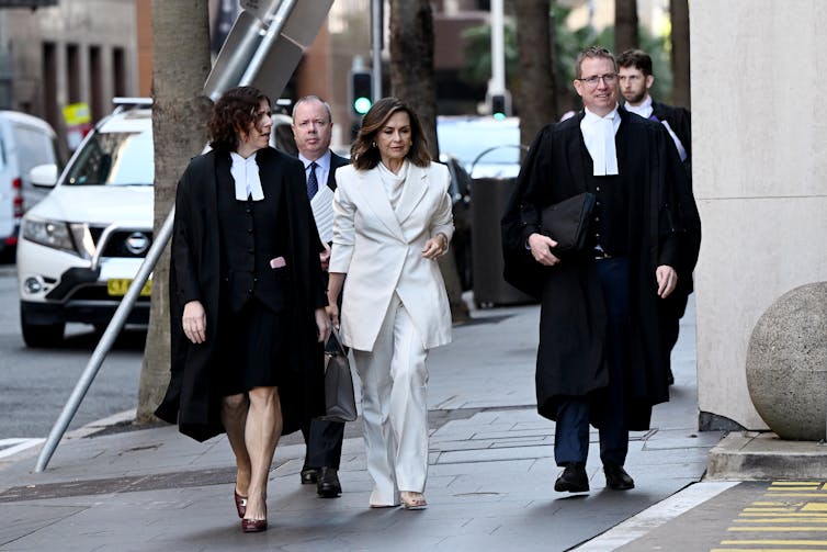 A woman in a white suit walks down the street in the middle of multiple lawyers in robes