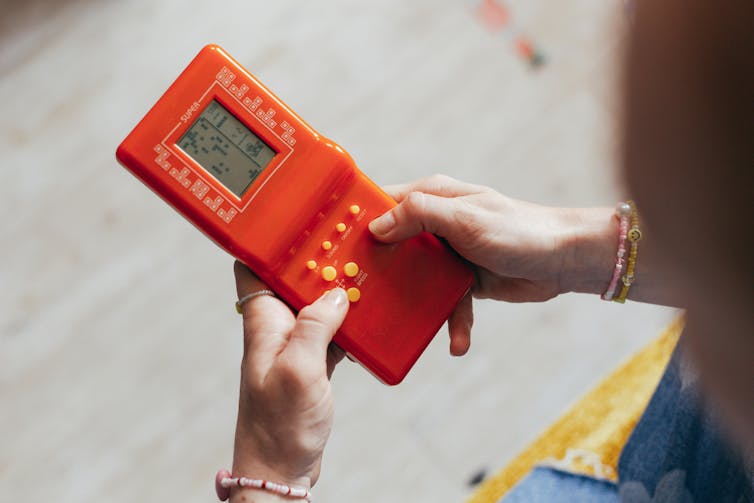 A person holding an orange retro Gameboy console and playing tetris on it.