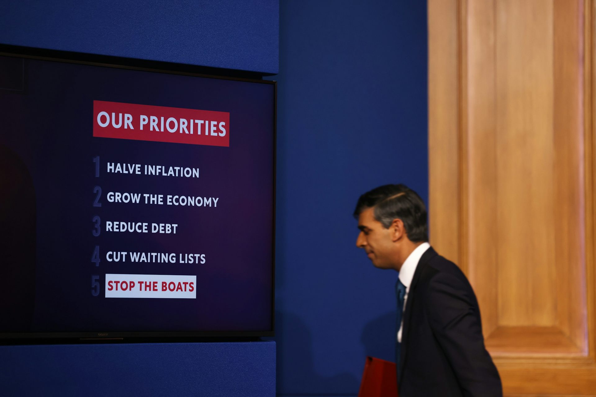 Side view of Rishi Sunak walking off a stage holding a red folder, there is a screen beside him listing the government's five priorities, with 'stop the boats' highlighted