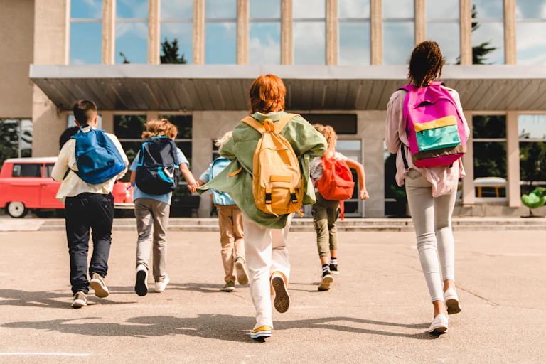 View from behind of six young students dressed in colourful clothing and backpacks, running into a school building