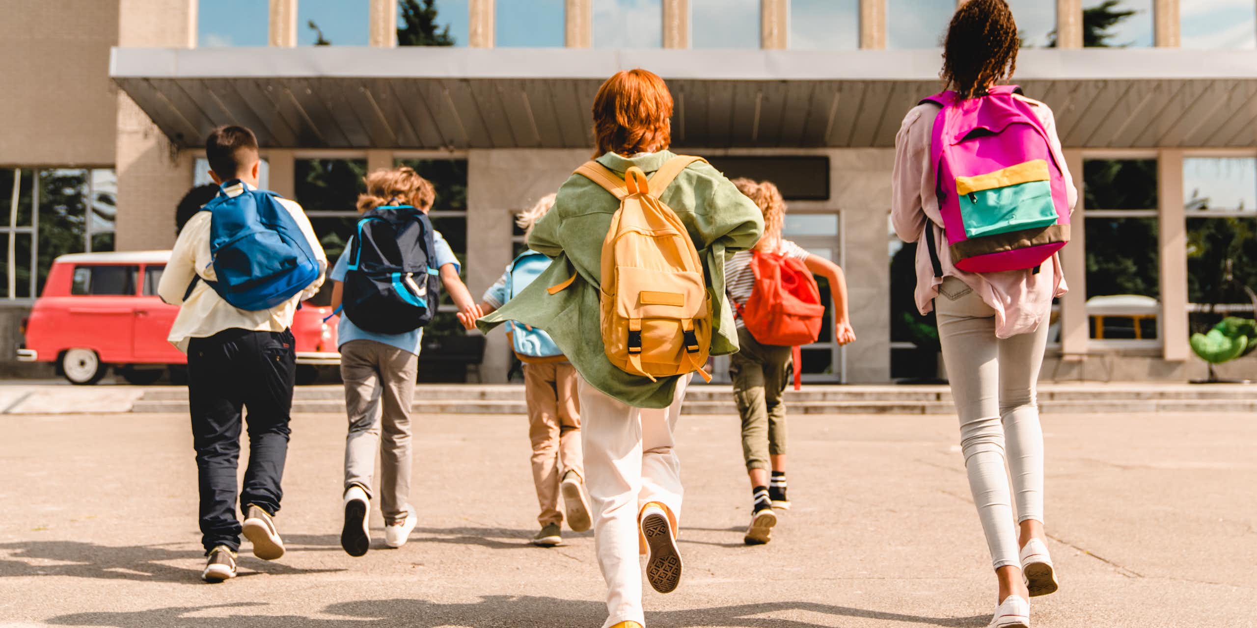 View from behind of six young students dressed in colourful clothing and backpacks, running into a school building
