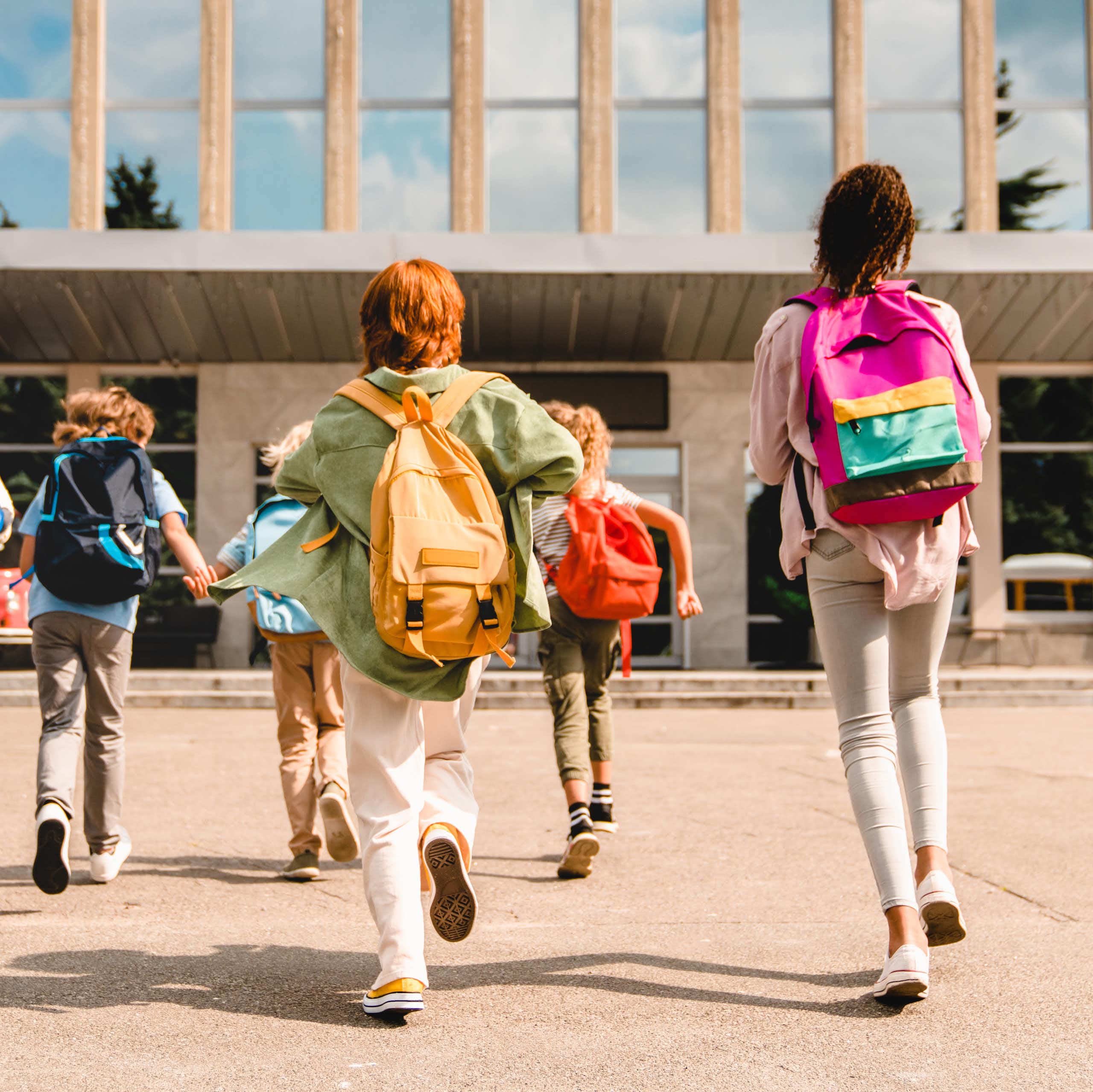 View from behind of six young students dressed in colourful clothing and backpacks, running into a school building