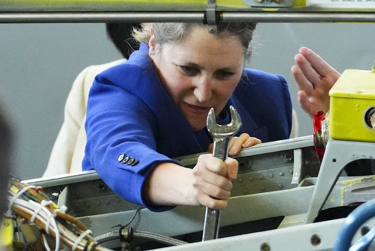 A woman in a blue jacket concentrates as she holds a wrench.