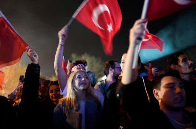 A woman in a crowd of people waving a red and white flag smiles as she looks towards a stage where someone is speaking.