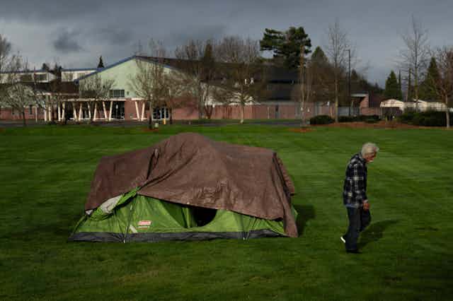 A man walks past a tent on a grassy field, with a school in the background