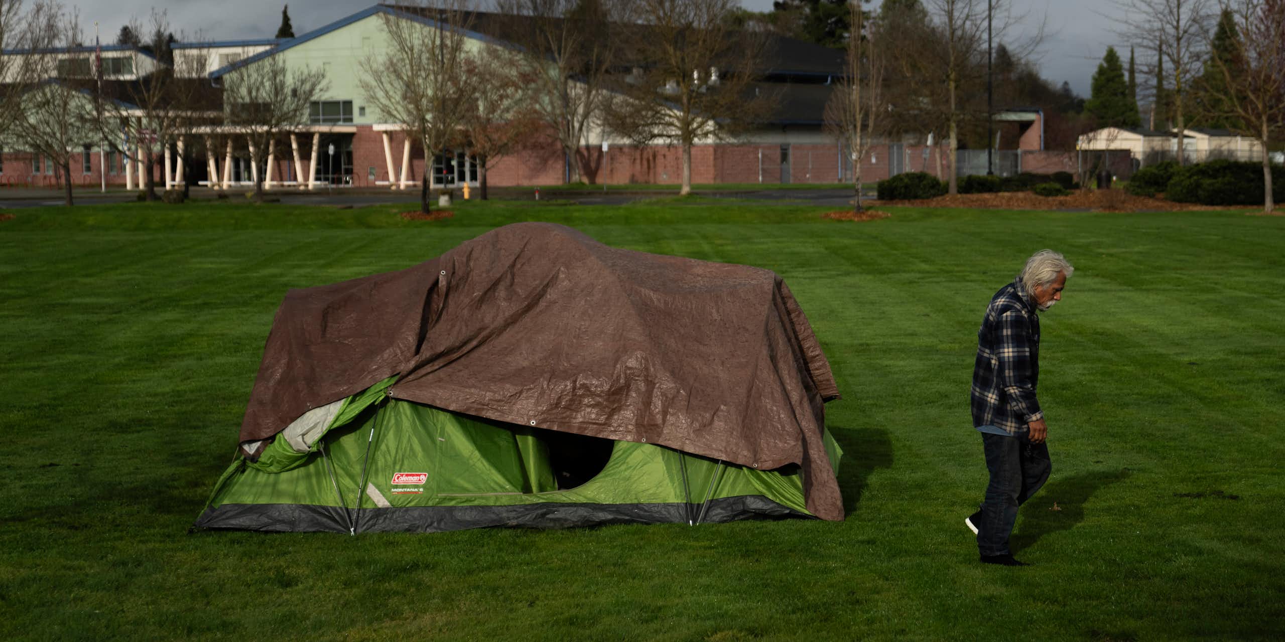 A man walks past a tent on a grassy field, with a school in the background