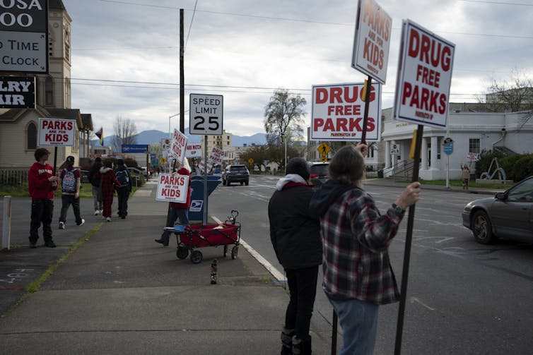 People stand on a sidewalk holding signs reading 'Parks Are for Kids' and 'Drug Free Parks'