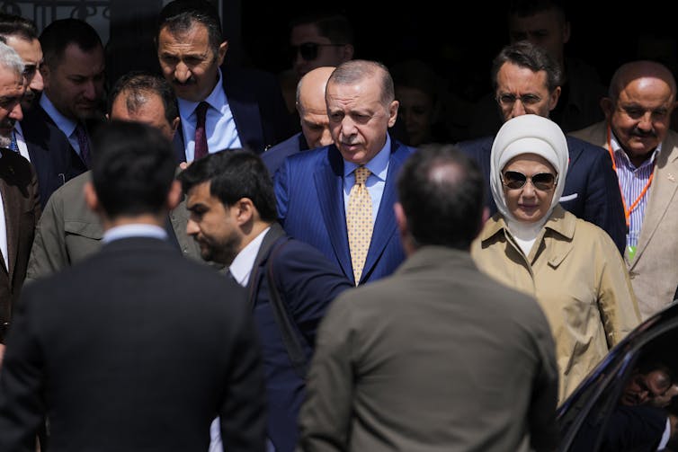 A man in a suit surrounded by other men leaves a polling station next to a woman with a white kerchief on her head wearing a trenchcoat.