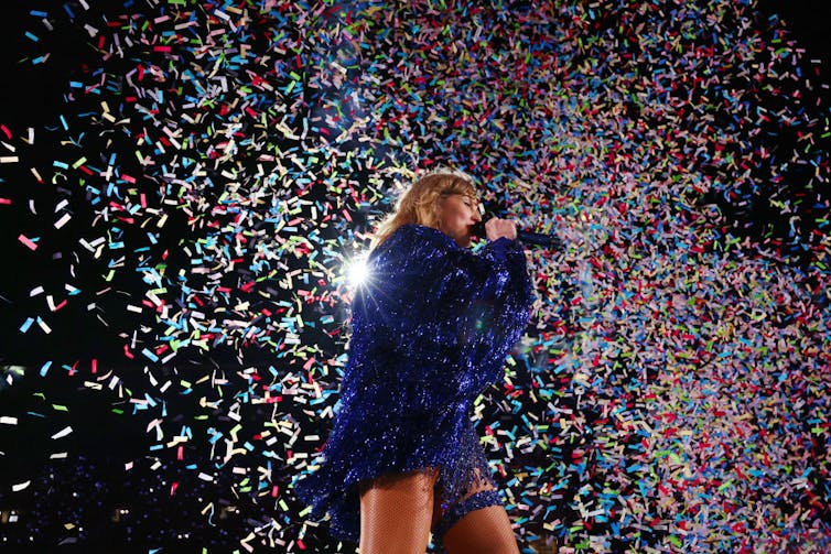 A blonde woman in a sparkly blue coat sings into a microphone as rainbow-colored confetti falls around her.